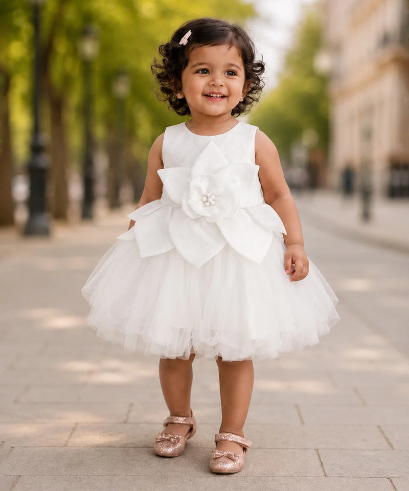 A toddler girl wearing a sleeveless white tulle dress with a large fabric flower applique on the front, standing on a paved outdoor walkway with blurred trees and buildings in the background.