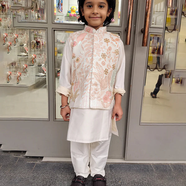 Child wearing a cream color kurta pajama with a jacket  traditional outfit standing in front of a store entrance.