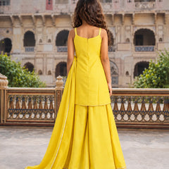 A woman stands with her back to the camera wearing a bright yellow traditional outfit with a long train, set against a historic stone courtyard with arches and greenery.