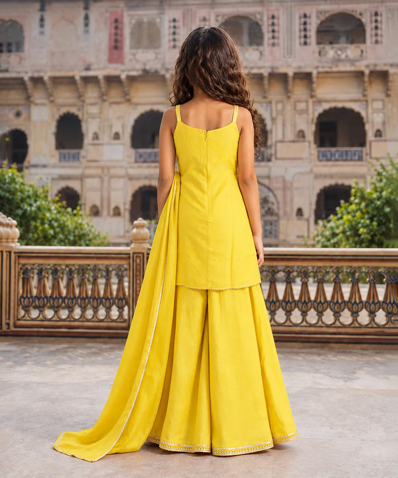 A woman stands with her back to the camera wearing a bright yellow traditional outfit with a long train, set against a historic stone courtyard with arches and greenery.