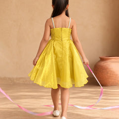 A young girl viewed from behind wearing a bright yellow sleeveless dress with thin white straps, standing in a warm beige indoor setting and holding a pink ribbon.