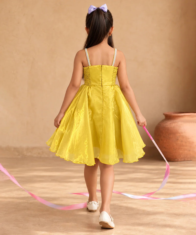 A young girl viewed from behind wearing a bright yellow sleeveless dress with thin white straps, standing in a warm beige indoor setting and holding a pink ribbon.