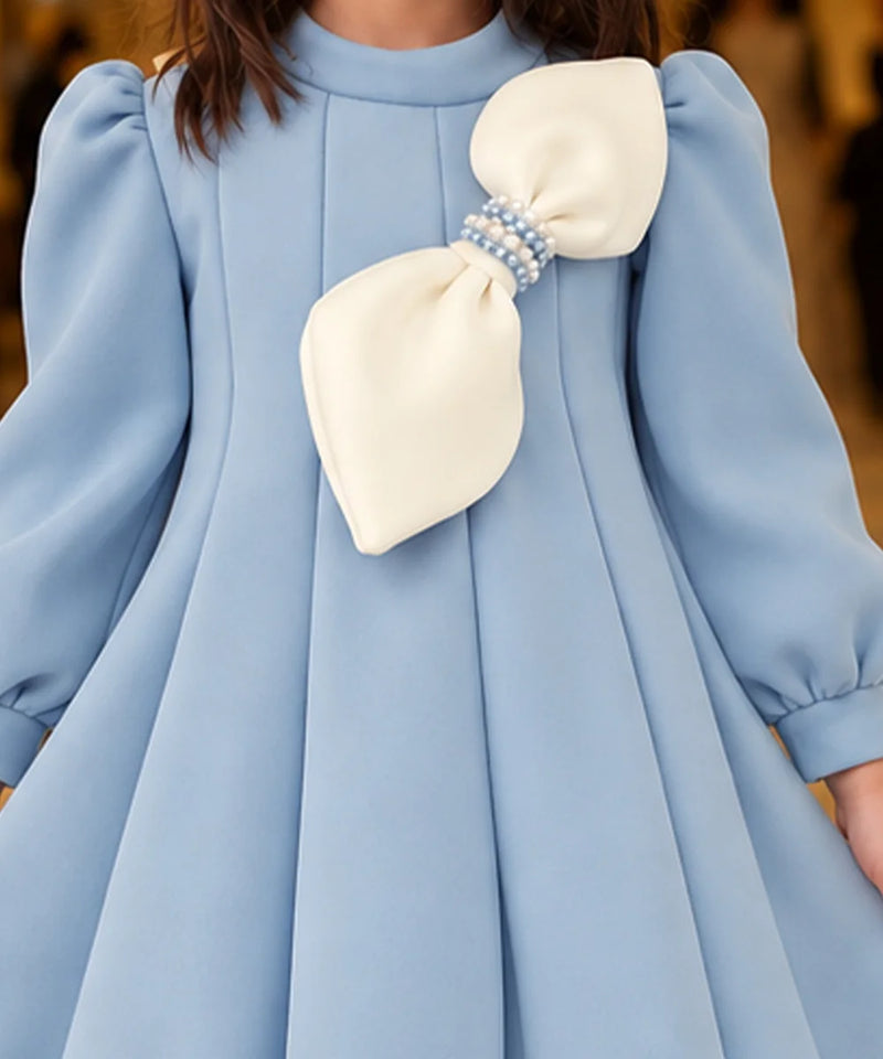 A close-up of a light blue dress with a large cream bow on the chest, featuring a pearl-like beaded band at the center of the bow.