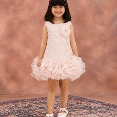 A young girl with dark hair wearing a sleeveless light pink dress with a textured bodice, a large fabric flower on the chest, and a layered ruffle skirt, standing on a patterned rug against a textured pink studio backdrop.