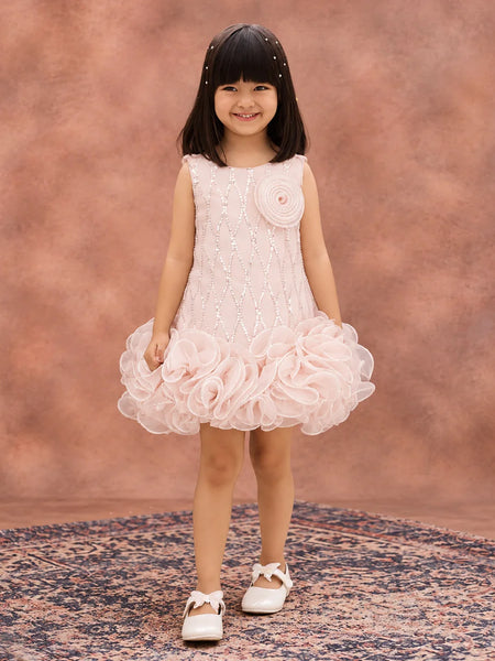 A young girl with dark hair wearing a sleeveless light pink dress with a textured bodice, a large fabric flower on the chest, and a layered ruffle skirt, standing on a patterned rug against a textured pink studio backdrop.
