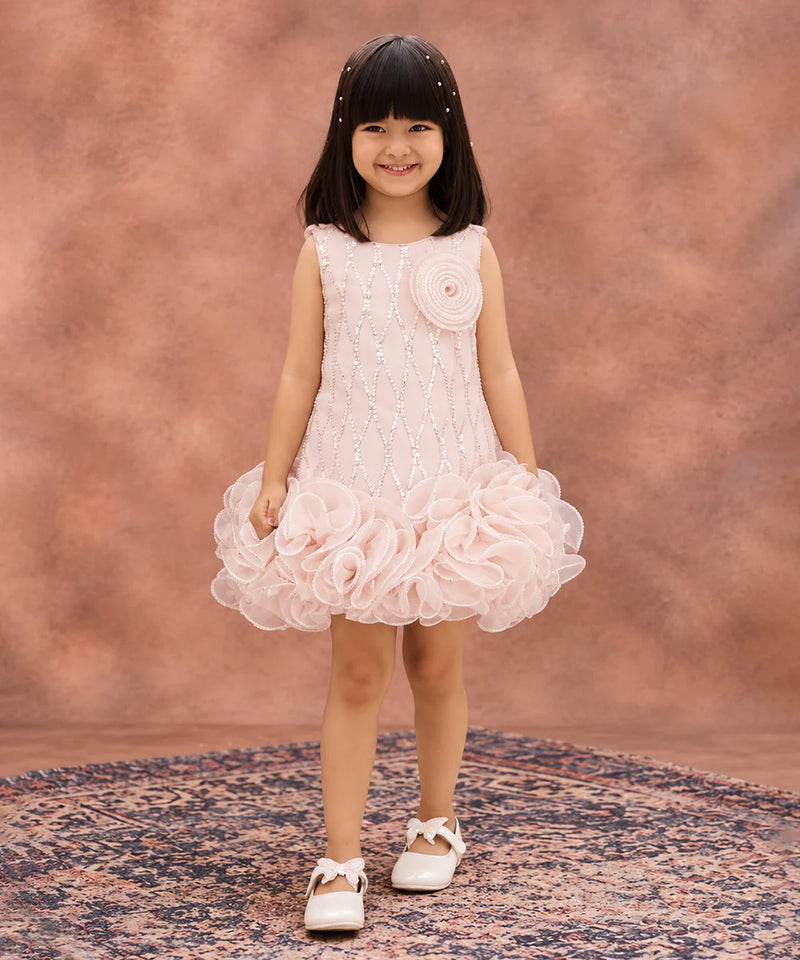 A young girl with dark hair wearing a sleeveless light pink dress with a textured bodice, a large fabric flower on the chest, and a layered ruffle skirt, standing on a patterned rug against a textured pink studio backdrop.