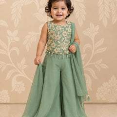 A toddler girl wearing a sage green traditional outfit with a floral embroidered bodice and a matching sheer dupatta, standing against a beige floral patterned wall.