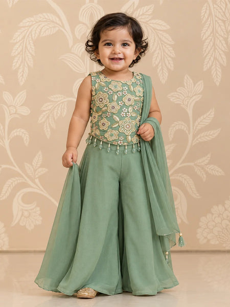 A toddler girl wearing a sage green traditional outfit with a floral embroidered bodice and a matching sheer dupatta, standing against a beige floral patterned wall.