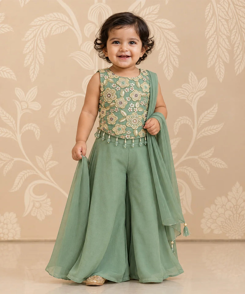 A toddler girl wearing a sage green traditional outfit with a floral embroidered bodice and a matching sheer dupatta, standing against a beige floral patterned wall.