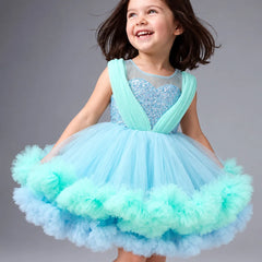 A young girl with dark hair is smiling and looking to the side while wearing a light blue and mint green tutu dress with a sparkly bodice, photographed against a solid gray studio background.