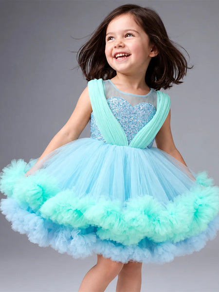 A young girl with dark hair is smiling and looking to the side while wearing a light blue and mint green tutu dress with a sparkly bodice, photographed against a solid gray studio background.