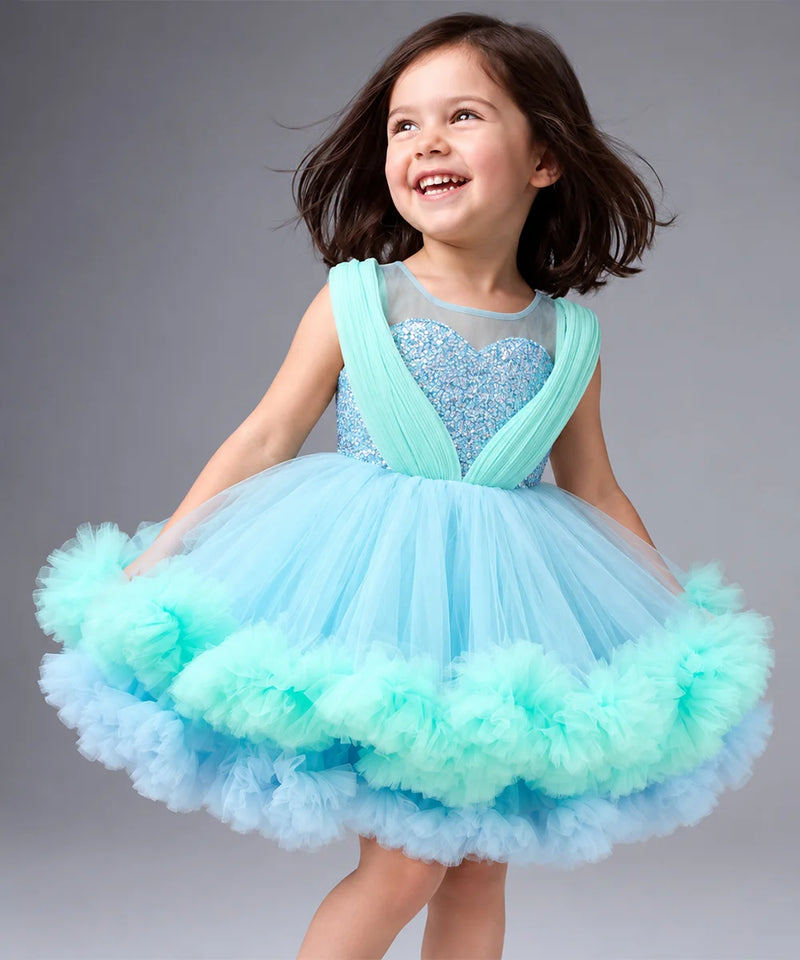 A young girl with dark hair is smiling and looking to the side while wearing a light blue and mint green tutu dress with a sparkly bodice, photographed against a solid gray studio background.