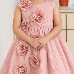 A young girl wearing a sleeveless dusty pink dress with large 3D fabric flowers on the bodice and skirt, standing indoors.