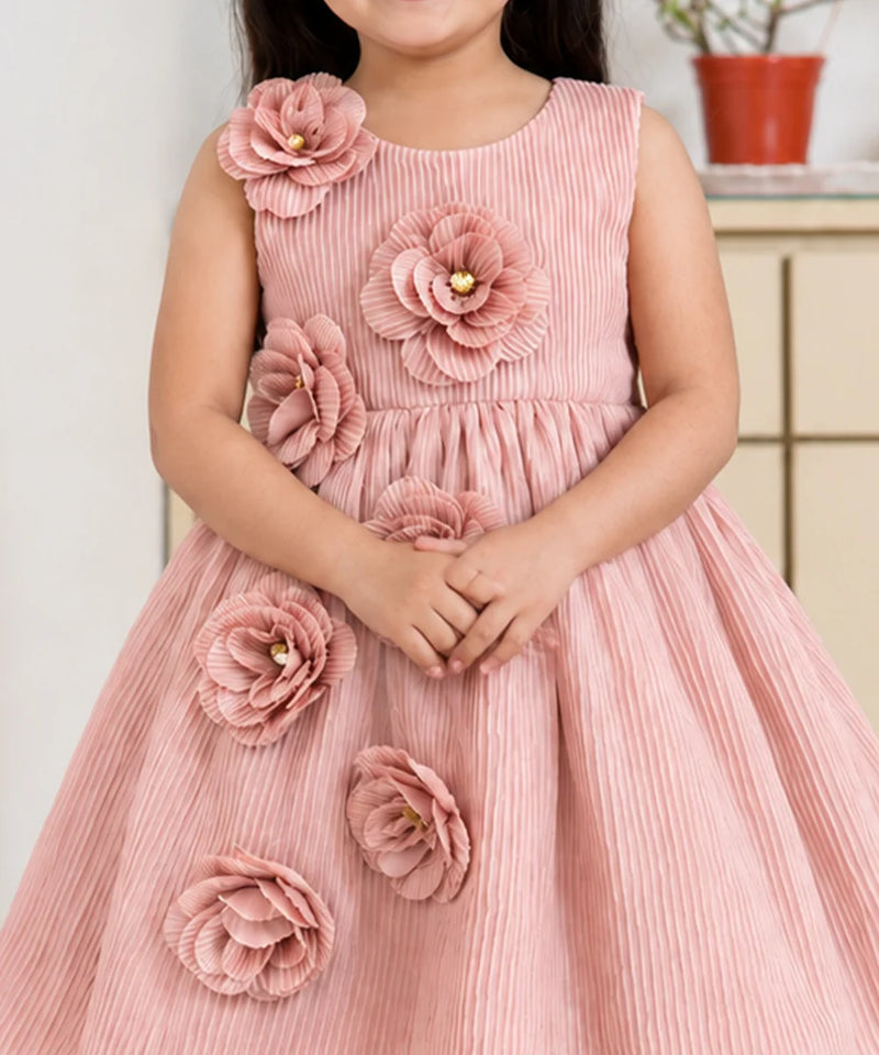 A young girl wearing a sleeveless dusty pink dress with large 3D fabric flowers on the bodice and skirt, standing indoors.