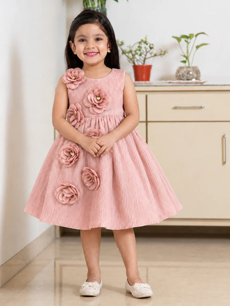 A young girl stands indoors wearing a sleeveless pink pleated dress with large fabric flowers attached to the bodice and skirt. The dress has a round neckline and a flared skirt, and she is wearing white shoes.