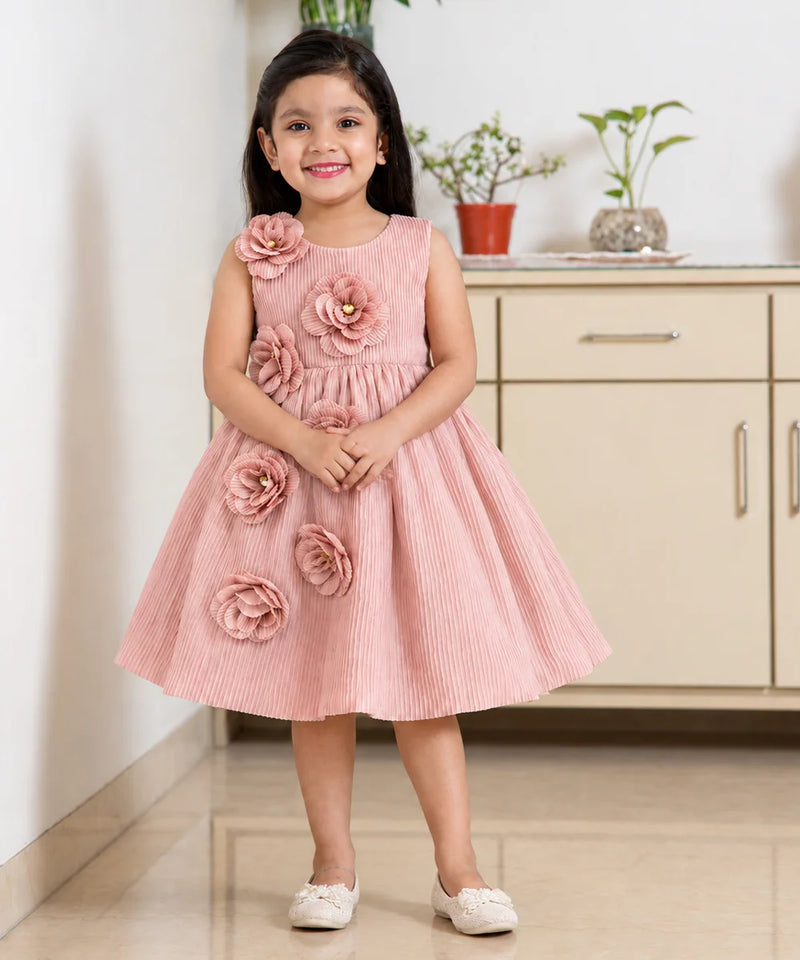 A young girl stands indoors wearing a sleeveless pink pleated dress with large fabric flowers attached to the bodice and skirt. The dress has a round neckline and a flared skirt, and she is wearing white shoes.