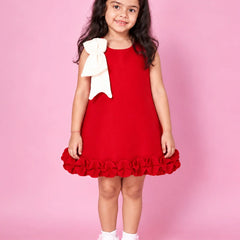 A young girl with long dark hair stands against a solid pink studio background wearing a sleeveless bright red dress with a large white bow on the shoulder and a ruffled hem, paired with white socks.