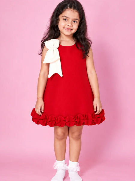 A young girl with long dark hair stands against a solid pink studio background wearing a sleeveless bright red dress with a large white bow on the shoulder and a ruffled hem, paired with white socks.
