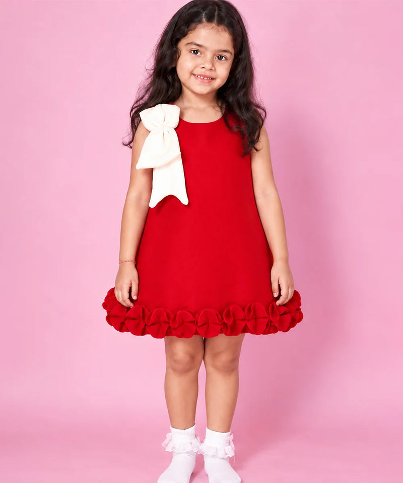 A young girl with long dark hair stands against a solid pink studio background wearing a sleeveless bright red dress with a large white bow on the shoulder and a ruffled hem, paired with white socks.