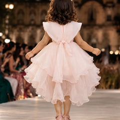 A young girl viewed from behind walking on a runway in a light pink sleeveless tulle dress with a ruffled tiered skirt and a bow at the back waist. The background shows a blurred indoor event space with warm lights and seated guests.