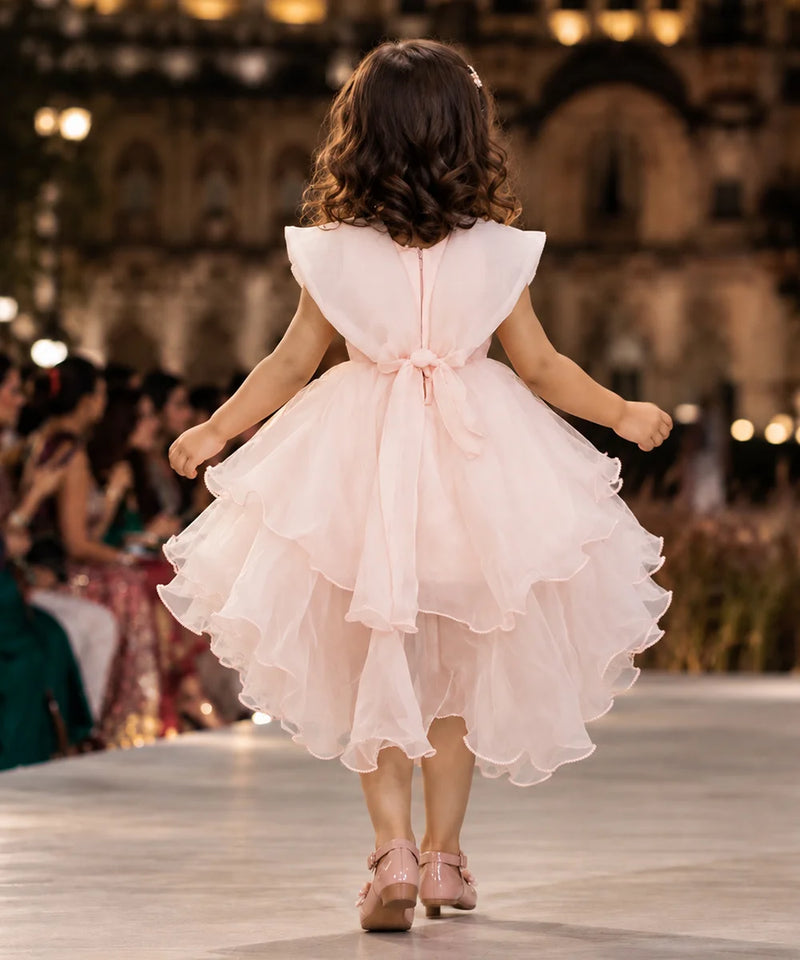 A young girl viewed from behind walking on a runway in a light pink sleeveless tulle dress with a ruffled tiered skirt and a bow at the back waist. The background shows a blurred indoor event space with warm lights and seated guests.