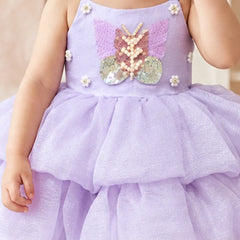 A toddler wearing a lavender purple sequined dress with a butterfly applique on the front, white floral accents, and a tiered tulle skirt.