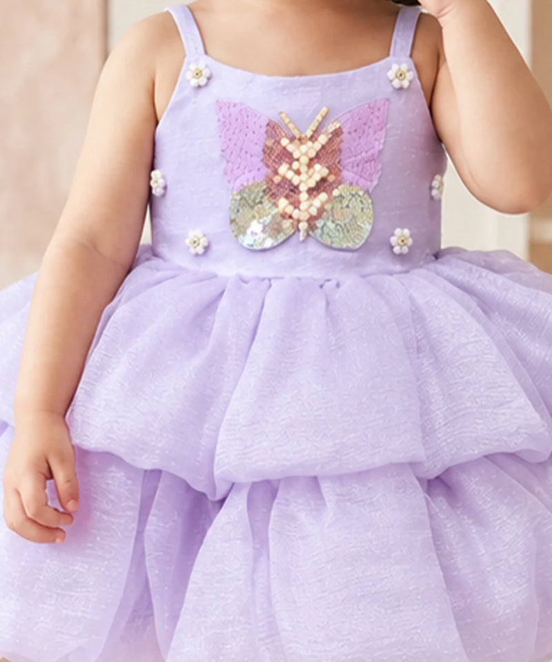 A toddler wearing a lavender purple sequined dress with a butterfly applique on the front, white floral accents, and a tiered tulle skirt.