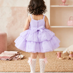A toddler girl viewed from behind wearing a lavender purple tiered tulle dress with a bow at the back, white lace socks, and white shoes, standing in a soft pink nursery setting.