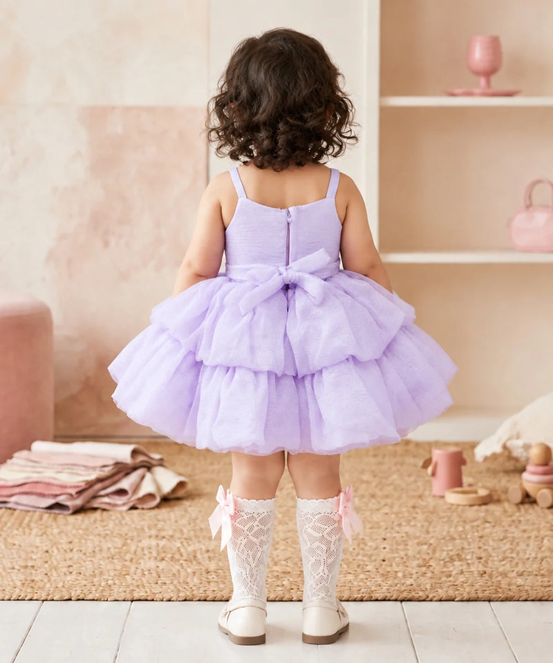 A toddler girl viewed from behind wearing a lavender purple tiered tulle dress with a bow at the back, white lace socks, and white shoes, standing in a soft pink nursery setting.