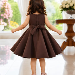 A young girl viewed from behind wearing a sleeveless dark brown A-line dress with a large bow at the back waist, standing in a bright indoor room with a tiled floor and floral decorations.