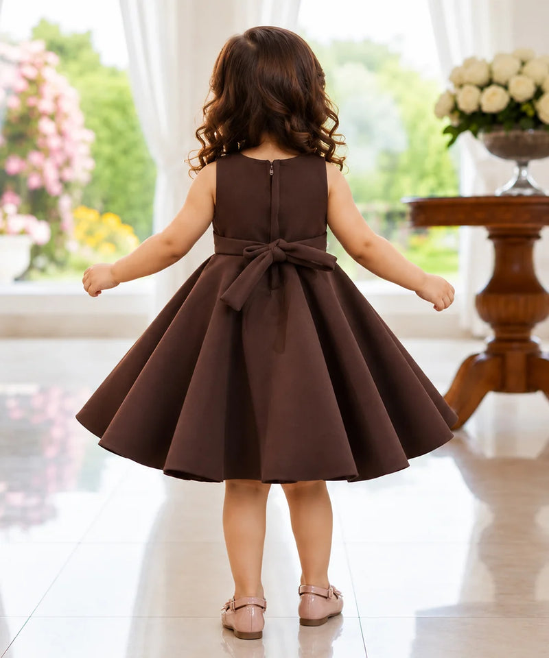 A young girl viewed from behind wearing a sleeveless dark brown A-line dress with a large bow at the back waist, standing in a bright indoor room with a tiled floor and floral decorations.