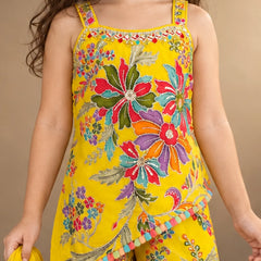 A child wearing a bright yellow sleeveless jumpsuit with colorful floral embroidery and multicolored pom-pom trim, shown against a plain beige studio background.