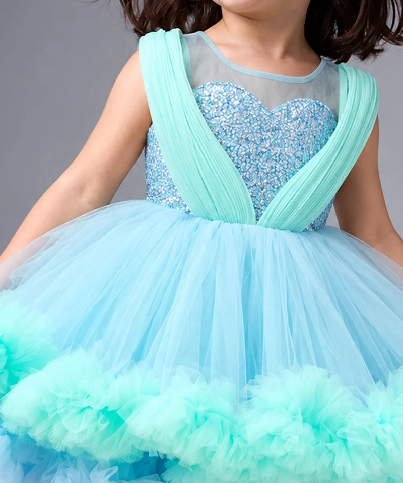 A child wearing a sleeveless light blue and mint green sequined ball gown with a sheer mesh bodice and a voluminous tulle skirt.