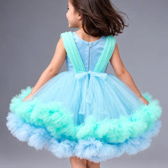 A young girl viewed from behind wearing a sleeveless light blue tutu dress with mint green ruffled trim and a bow at the back, photographed against a plain gray studio background.