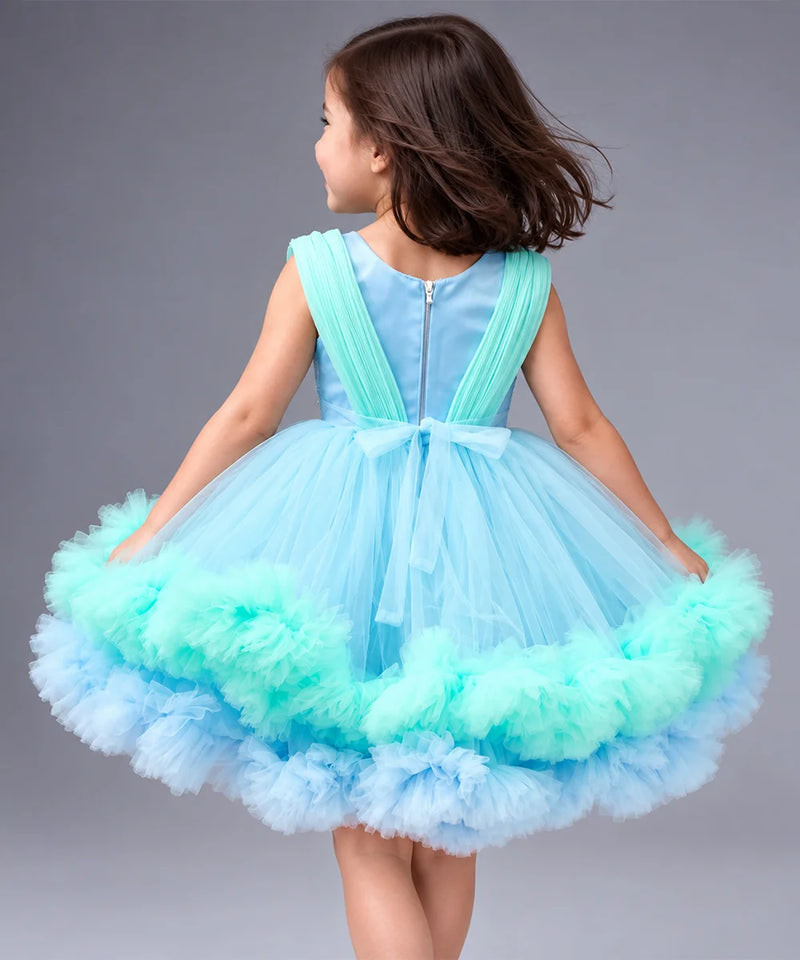 A young girl viewed from behind wearing a sleeveless light blue tutu dress with mint green ruffled trim and a bow at the back, photographed against a plain gray studio background.