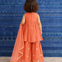 A toddler girl viewed from behind wearing a sleeveless coral-orange traditional outfit with matching wide-leg pants and a sheer dupatta, standing against a blue patterned backdrop.