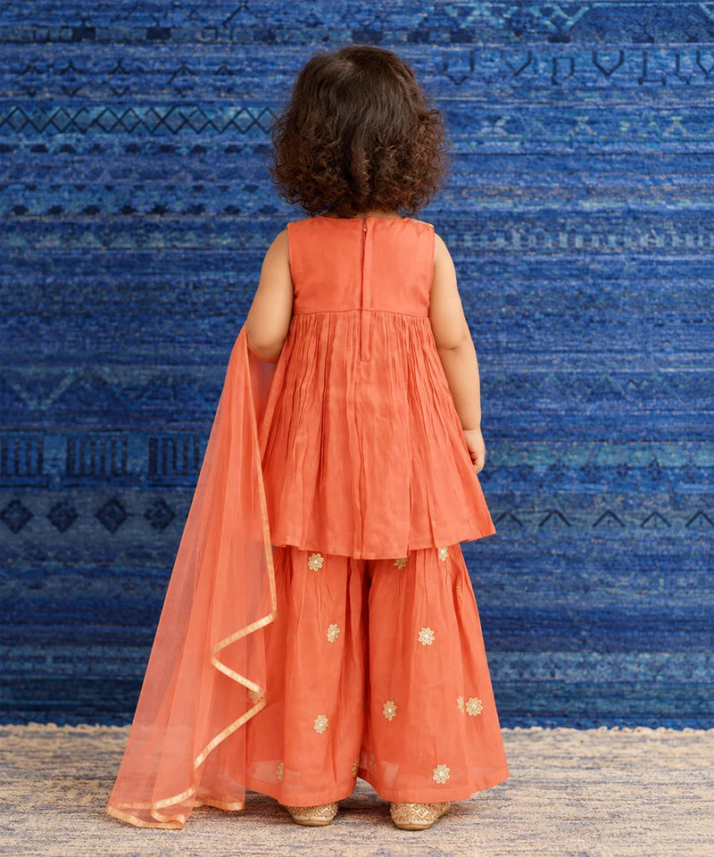 A toddler girl viewed from behind wearing a sleeveless coral-orange traditional outfit with matching wide-leg pants and a sheer dupatta, standing against a blue patterned backdrop.
