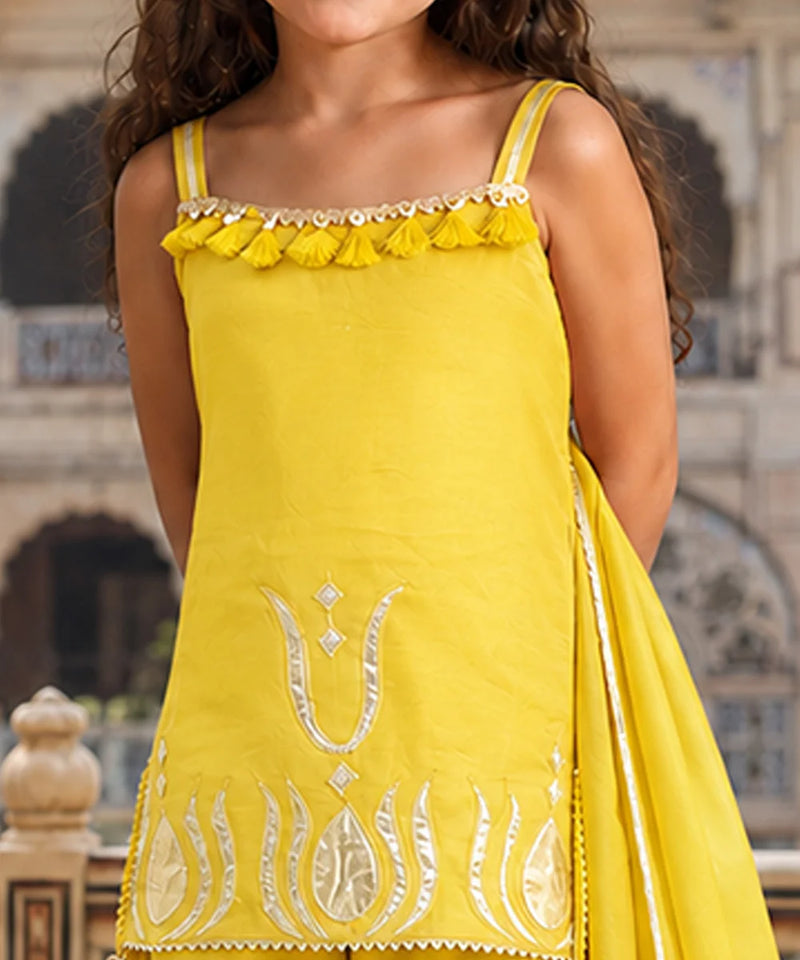 A bright yellow sleeveless traditional outfit with silver embroidery and tassel trim, worn by a model in front of a blurred architectural background.