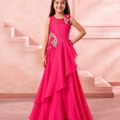 A young girl modeling a bright pink formal gown with silver embroidery and a layered ruffled skirt, standing in front of a pink architectural background.