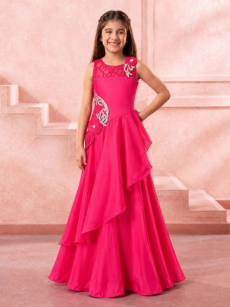 A young girl modeling a bright pink formal gown with silver embroidery and a layered ruffled skirt, standing in front of a pink architectural background.