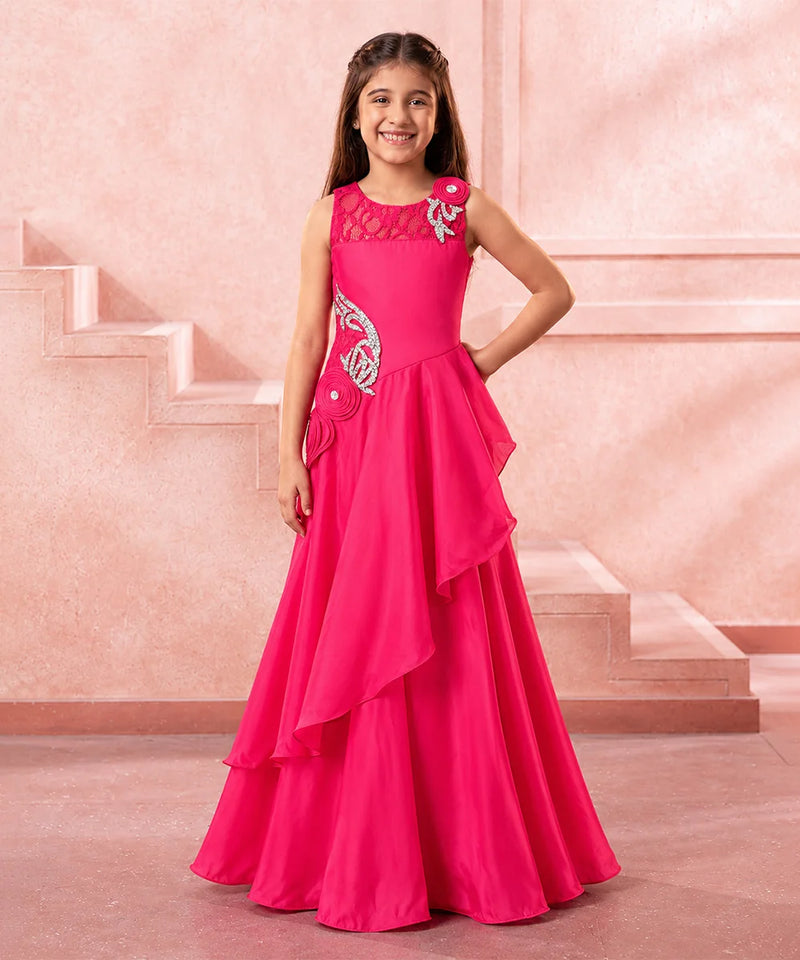 A young girl modeling a bright pink formal gown with silver embroidery and a layered ruffled skirt, standing in front of a pink architectural background.