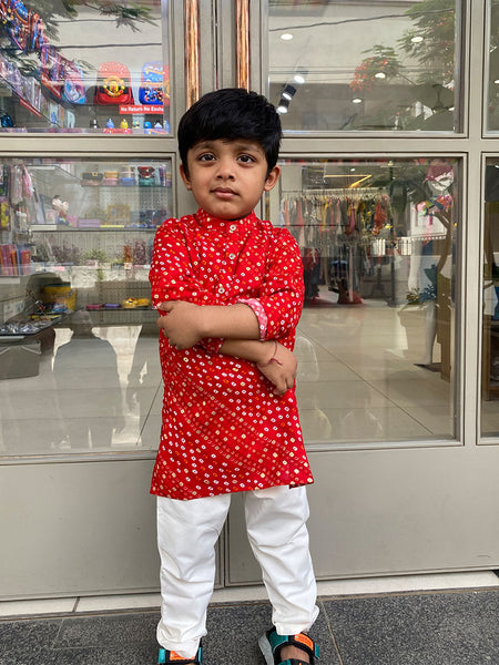 A young boy standing with his arms crossed wearing a red and white kurta pyjama set with a geometric pattern.