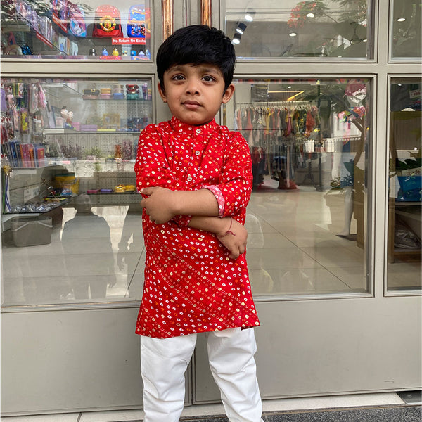 A young boy standing with his arms crossed wearing a red and white kurta pyjama set with a geometric pattern.