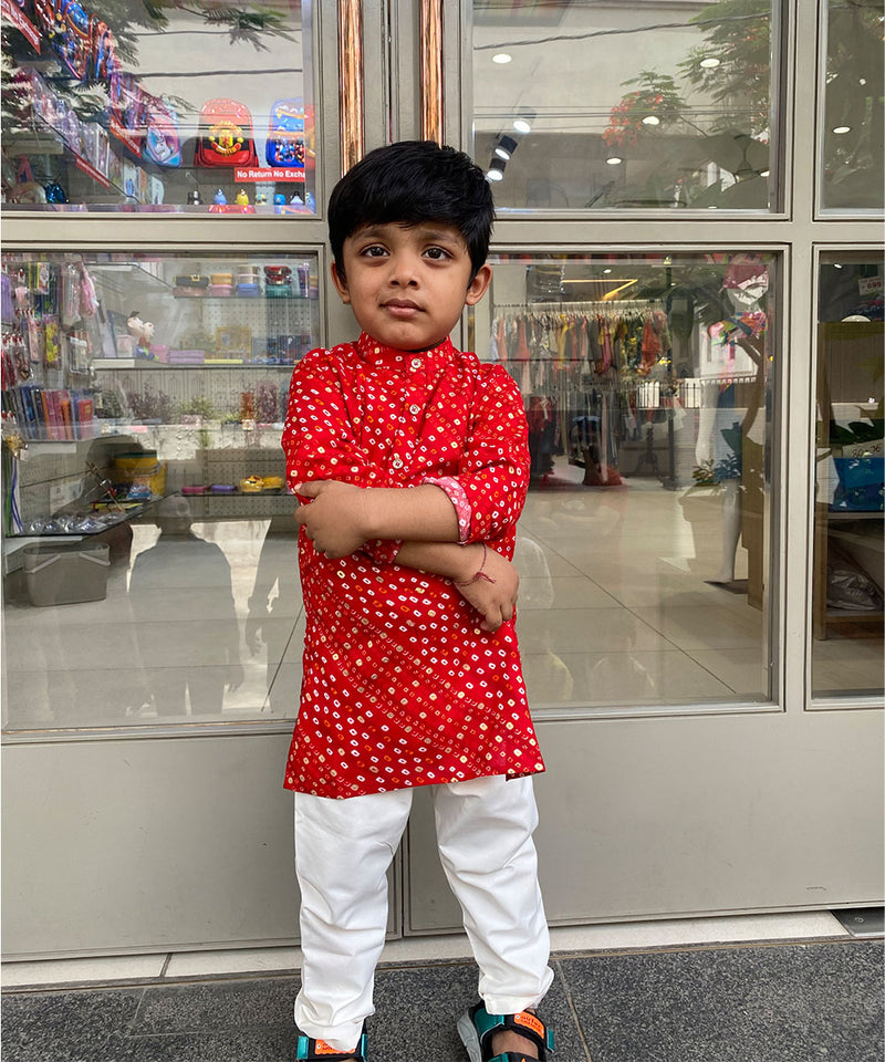 A young boy standing with his arms crossed wearing a red and white kurta pyjama set with a geometric pattern.