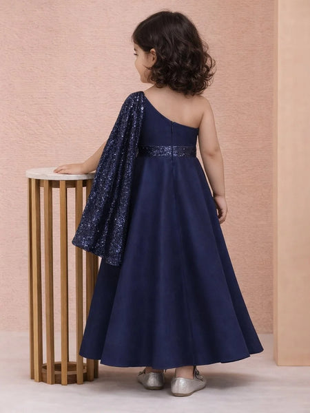 Child wearing a navy blue one-shoulder dress with sequin details, standing against a pink wall.