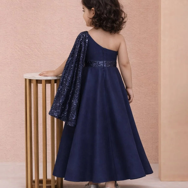 Child wearing a navy blue one-shoulder dress with sequin details, standing against a pink wall.