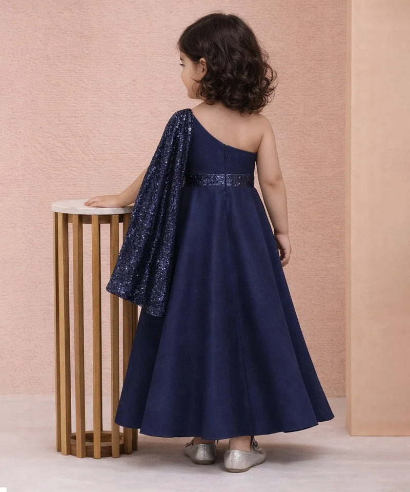 Child wearing a navy blue one-shoulder dress with sequin details, standing against a pink wall.