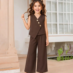 A young girl with curly brown hair wearing a dark brown two-piece outfit consisting of a sleeveless wrap vest with gold buttons and matching wide-leg trousers, paired with light blue shoes, standing in a classic indoor setting with a window and columns.