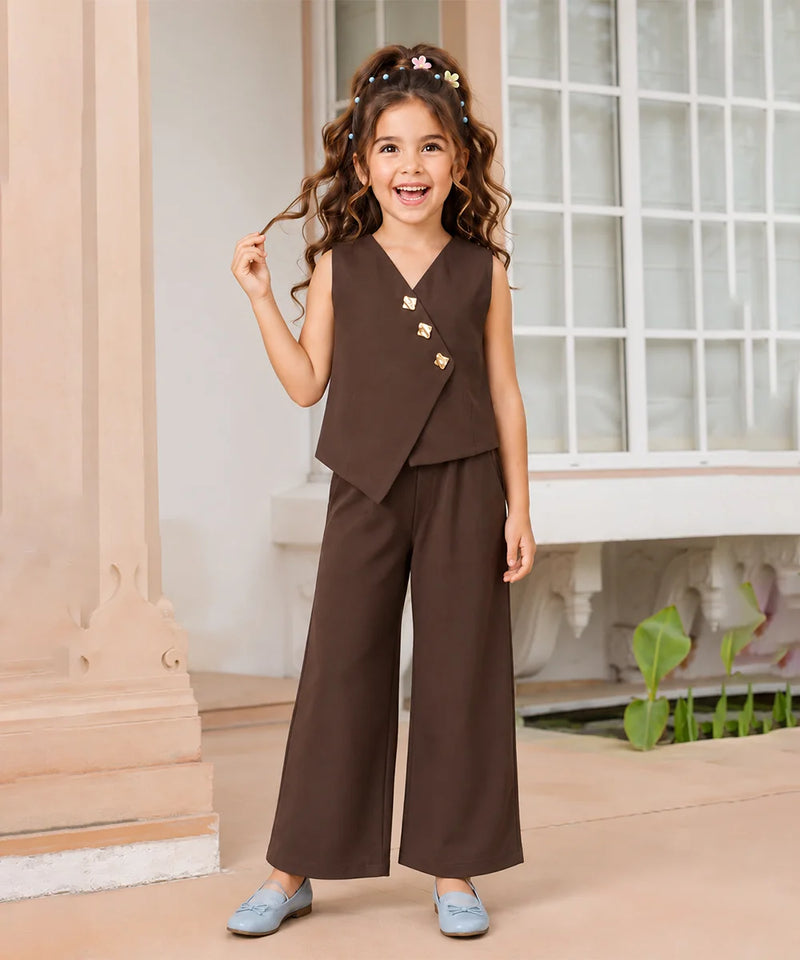 A young girl with curly brown hair wearing a dark brown two-piece outfit consisting of a sleeveless wrap vest with gold buttons and matching wide-leg trousers, paired with light blue shoes, standing in a classic indoor setting with a window and columns.