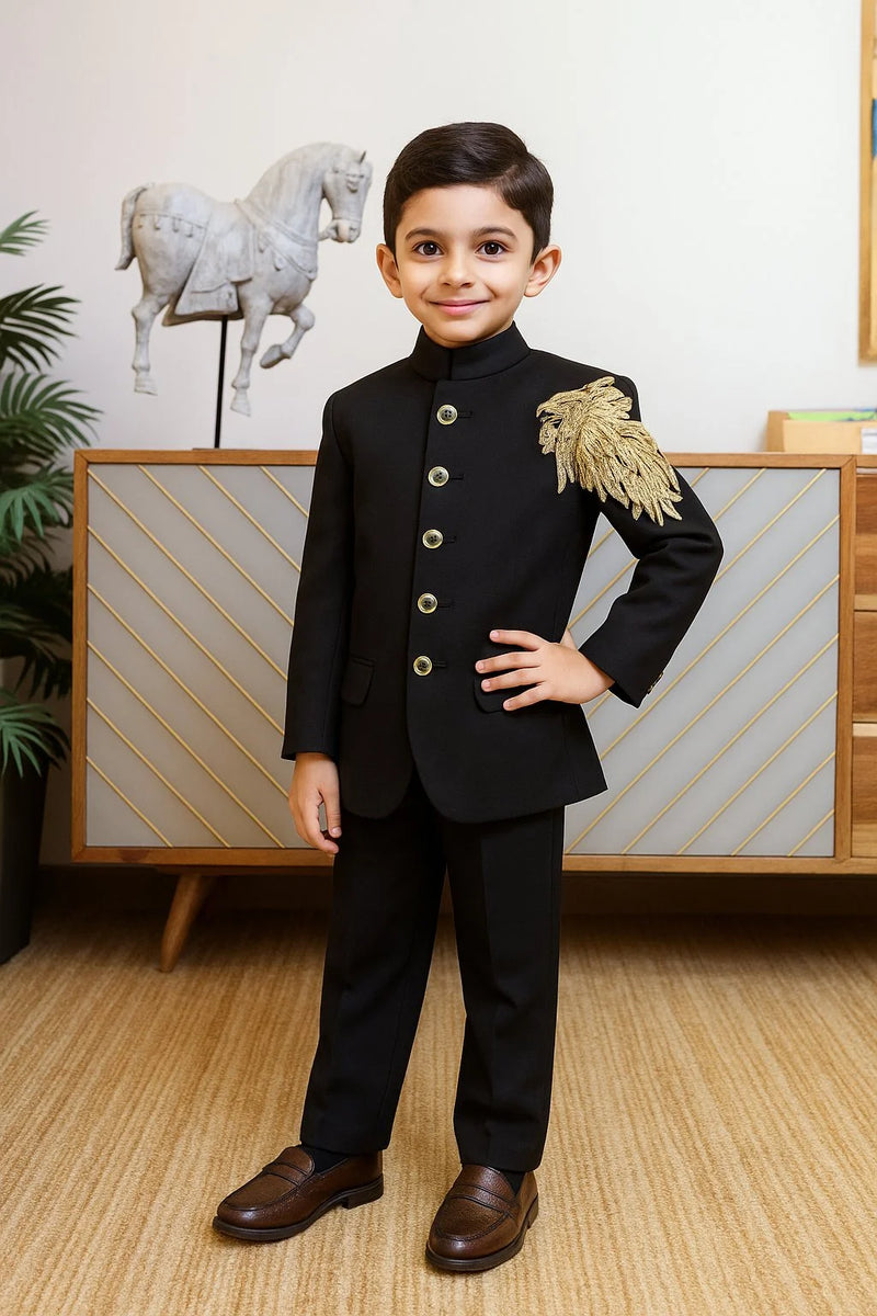 Young boy in a formal black suit with gold accents standing in a room with decorative elements.